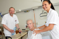 Politiker Axel Schäfer schenkt Patient Siegfried Sawatzki ein Glas Wasser ein, während Gesundheits- und Krankenpflegerin Jessica Gargulinski diesen stützt.